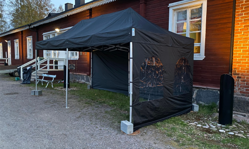 Musta katettu pop-up ulkoteltta, jossa on läpinäkyvät ikkunat sivuilla, sijoitettu nurmikolle puutalon ja rantamaiseman läheisyyteen.